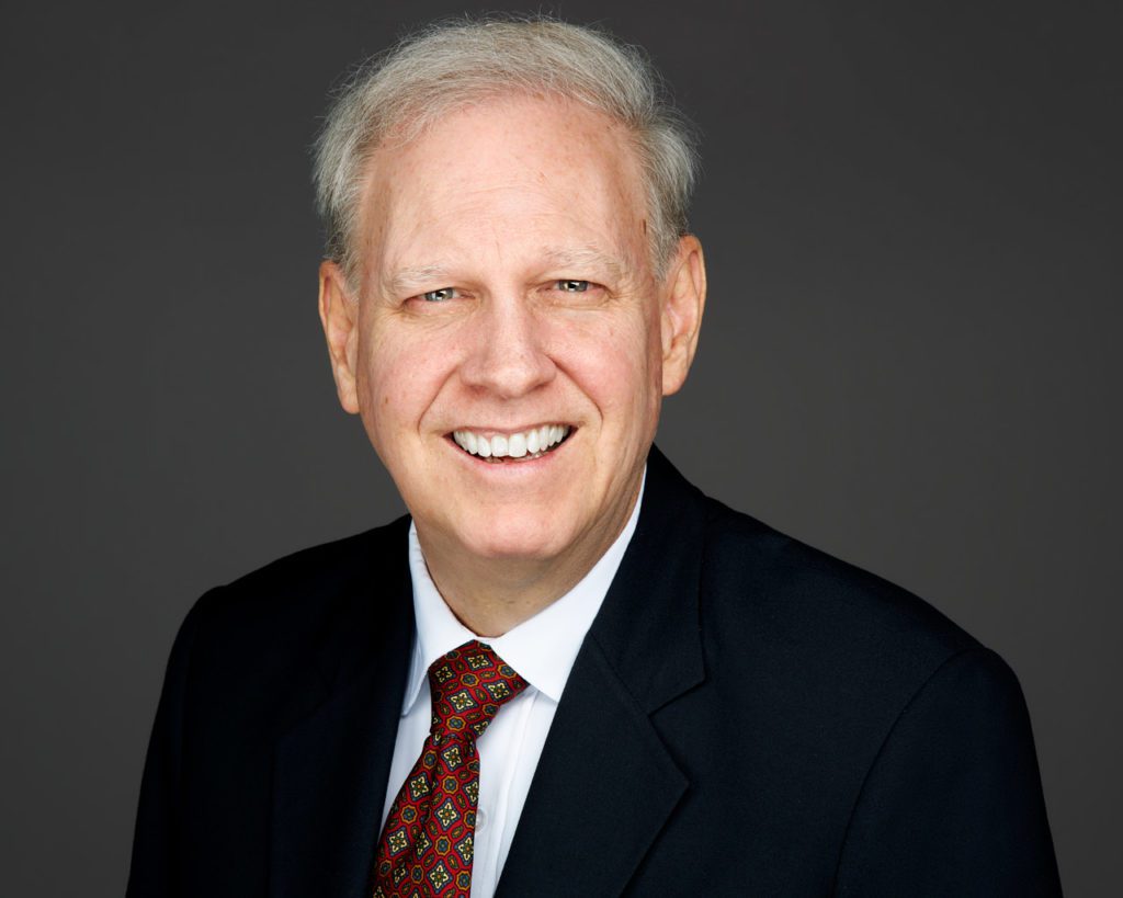 A photo of Dion Sullivan wearing a suit and tie smiling for a studio protrait.